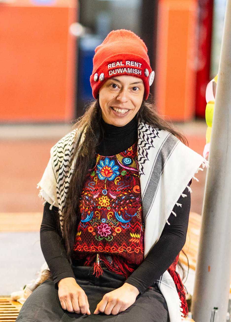Saunatina Sanchez, candidate for Seattle City Council Position 8 sits on a bench at a transit station.  She is wearing a red Real Rent Duwamish beanie over her long dark hair.  She is smiling into the camera.  He is wearing a floral embroidered shirt .