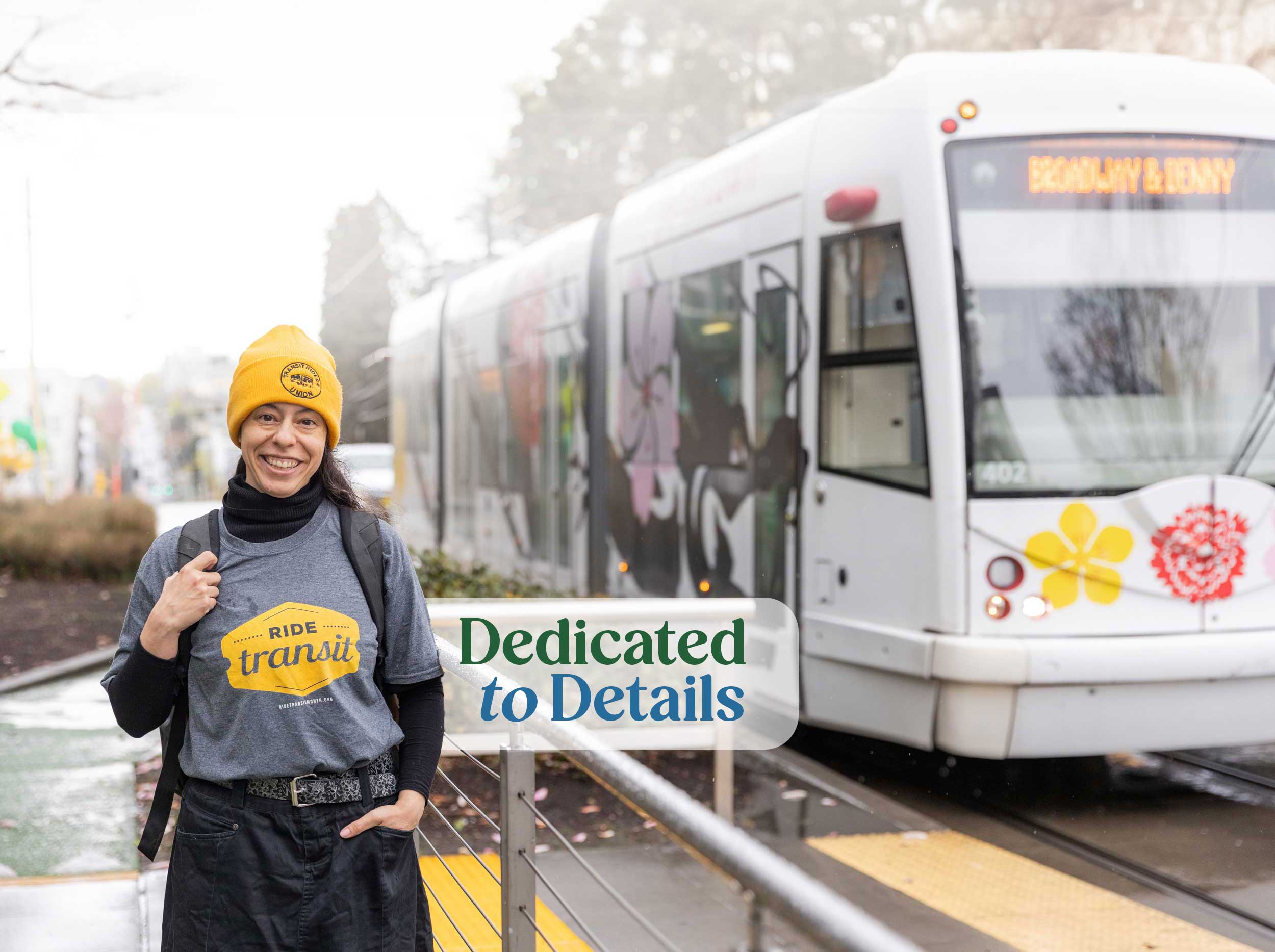 Saunatina Sanchez, candidate for Seattle City Council Position 6 stands in front of the Seattle Transit bus bound for Broadway & Denny.  She has long dark hair and dark eyes and is smiling.  She is wearing a Tranist Riders Union yellow colored beanie hat and a gray tshirt with a yellow Ride Tranist logo on it.
