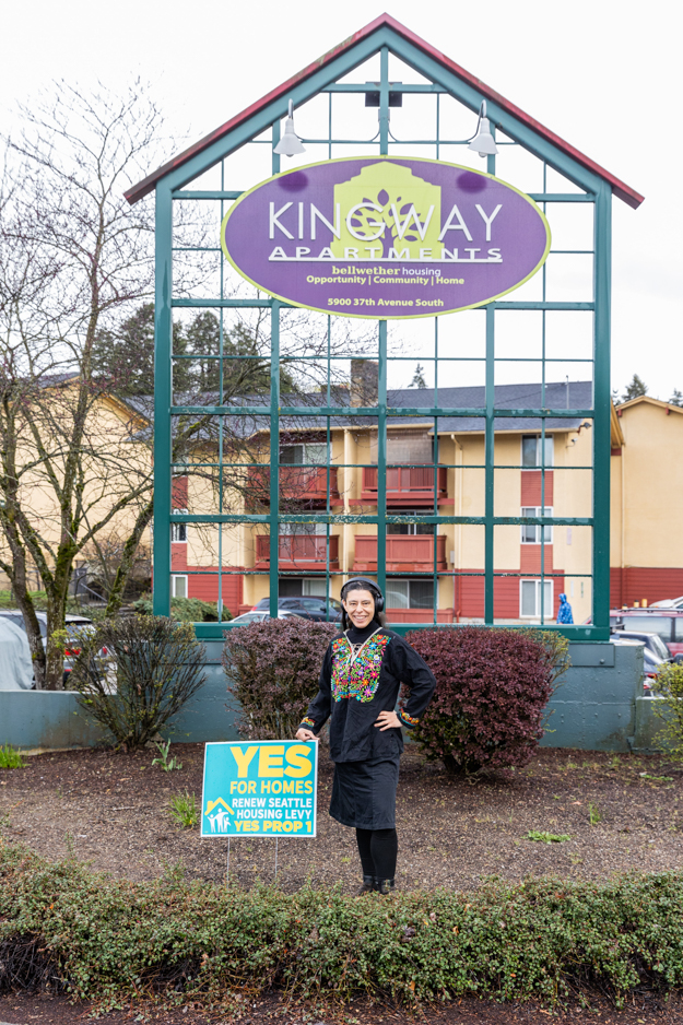 Saunatina Sanchez, candidate for Seattle City Council Position 8 stands in front of the Kingway Apartments and next to a sign that says Yes for Homes.  She is wearing a floral embroidered shirt and is smiling.