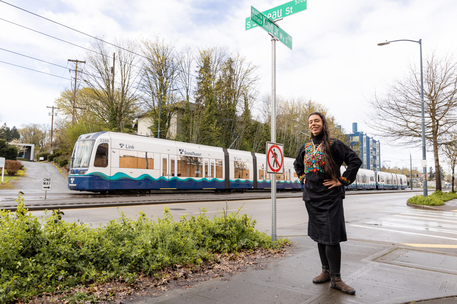 Saunatina Sanchez, candidate for Seattle City Council Position 8 stands in front of an intersection and a street sign.  A SEattle transit train is passing in the background.