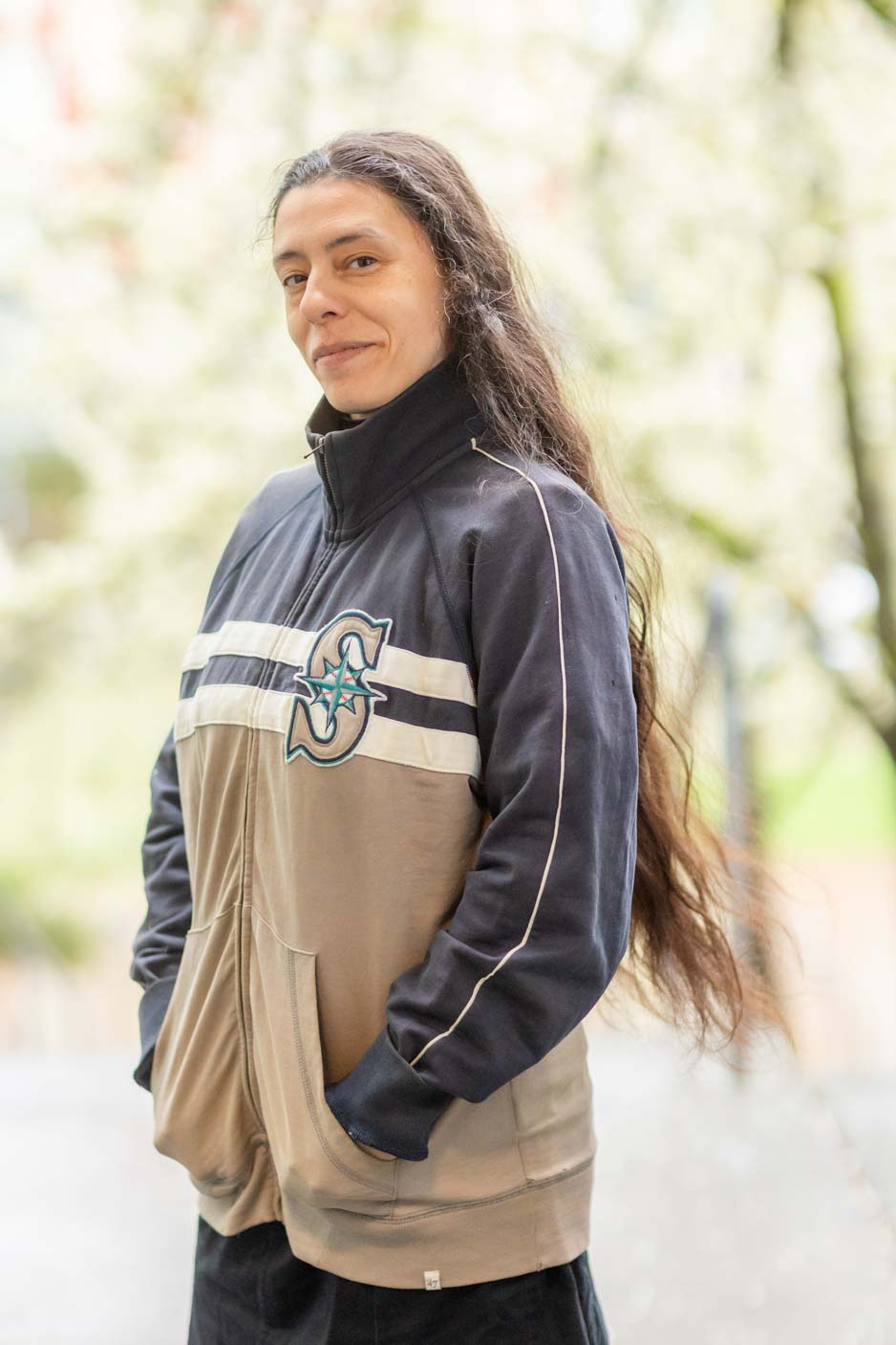 Saunatina Sanchez, candidate for Seattle City Council Position 8 is wearing a Seattle Mariners jacket with her hands in her pockets.  She has long dark hair and dark eyes and is looking directly at the camera.  
