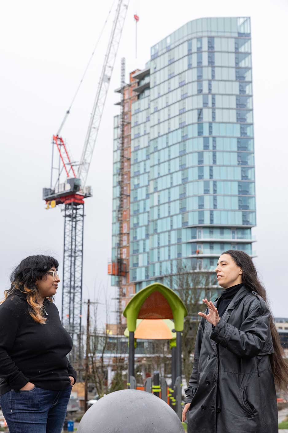 Saunatina Sanchez, candidate for Seattle City Council Position 8 has long dark hair and dark eyes.  She is speaking to another woman int front of a playground with a skyscaper and crane in the background.