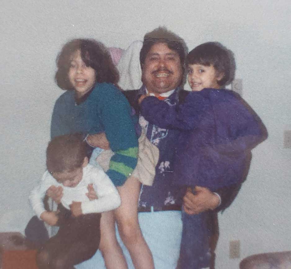Saunatina Sanchez and her siblings are being held up by her father in this family photo.  The father is standing strong and proud with a smile.  He has dark hair and a moustache and is smiling.  He has one child on each hip.  The child on the left is holding an even smaller child and they are all smiling.